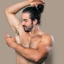 A young and muscular man with a man bun and no shirt topless applying pit spritz deodorizing spray to his armpit underarm against a gray background