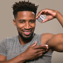 african american black man applying stank stop natural deodorant scotch pine and coriander to his armpit underarm against a gray backgrounf